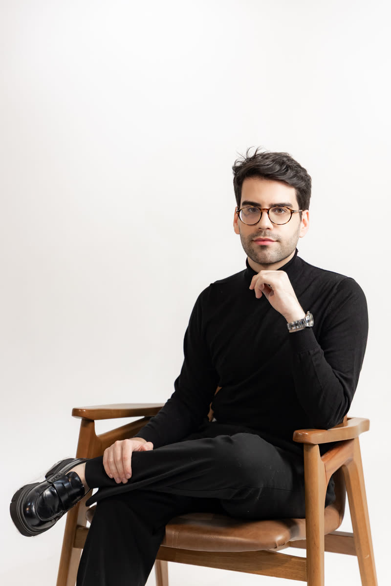 Lucas Bulcão sentado, de preto, em retrato de estúdio